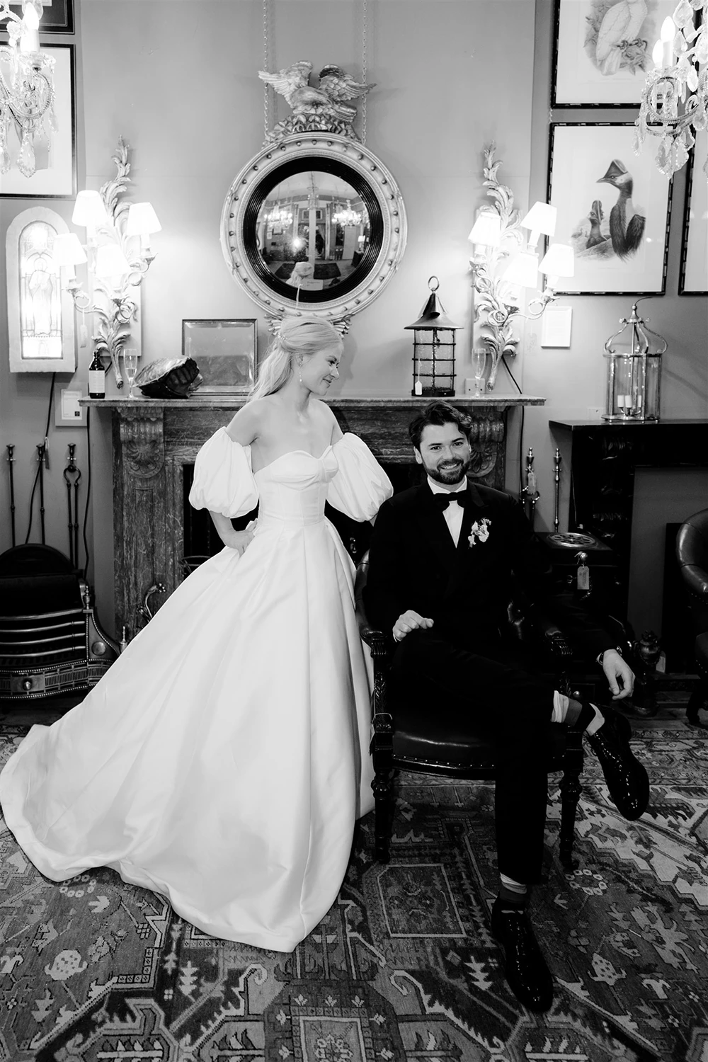 Black and white portrait of bride standing beside groom seated in an armchair in front of a decorative fireplace.
