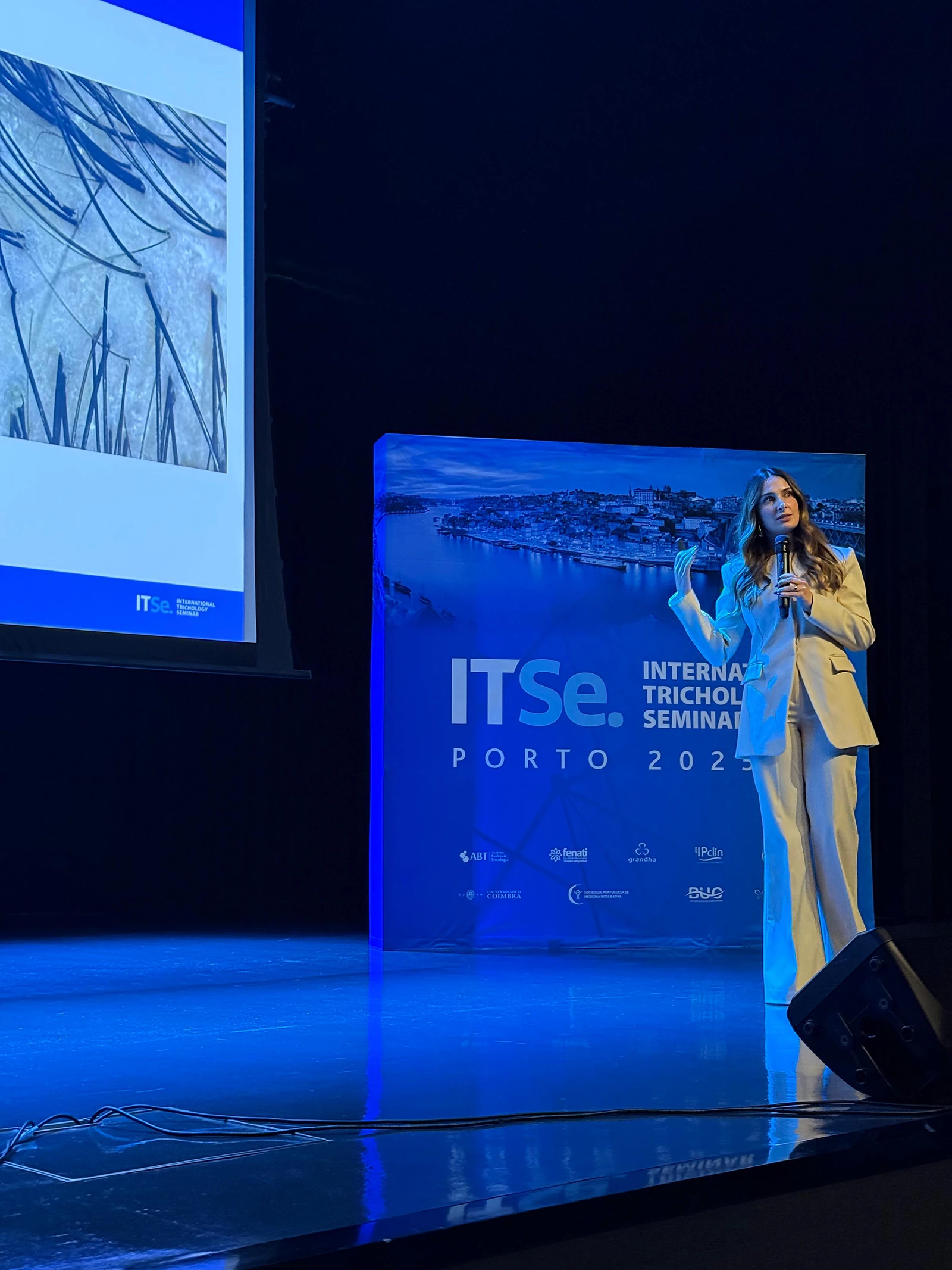 Izabella Bordignon speaks into a microphone while standing on stage next to an ITSe International Trichology Seminar Porto event backdrop banner.
