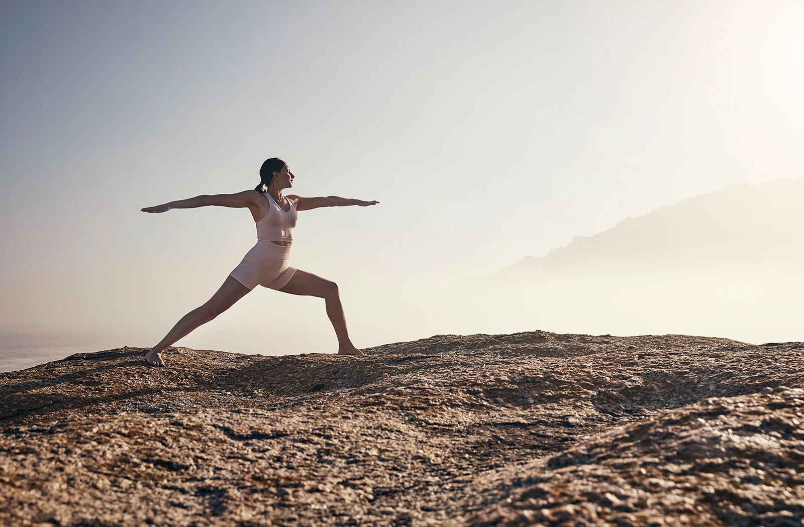 A woman practices yoga, balancing in a warrior pose on a rugged rocky surface outdoors against a misty background.