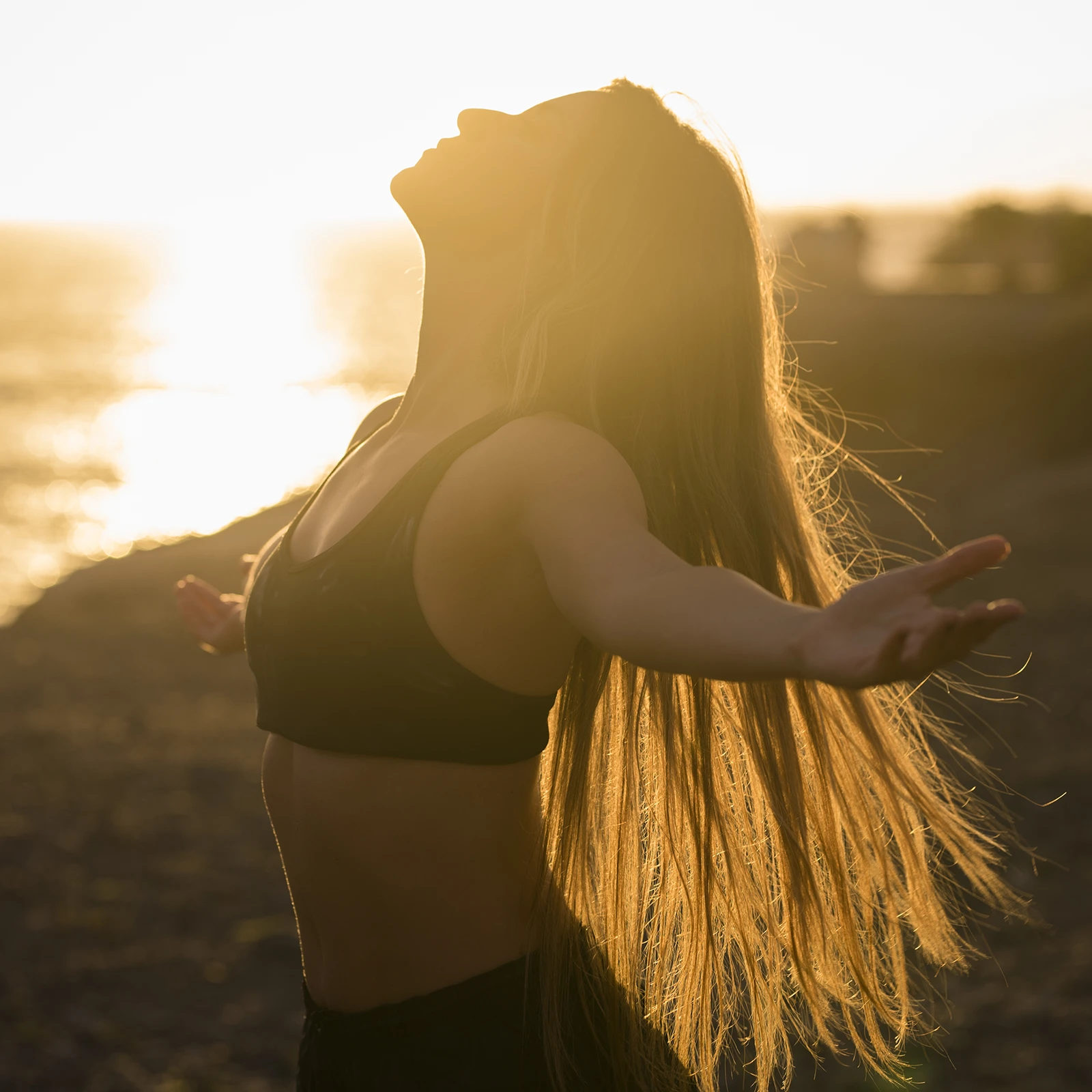 A woman with long hair stands outdoors at sunset with her arms outstretched, embracing the warm golden light in a peaceful moment that represents wellness, mindfulness, freedom, and connection with nature.