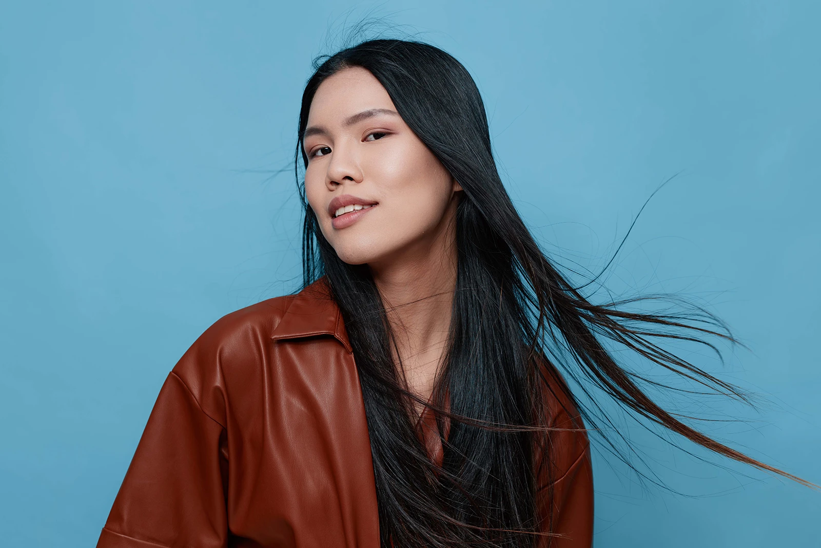 Studio portrait of a smiling Asian woman with long black hair blowing in the wind, wearing a brown leather jacket against a neutral background.