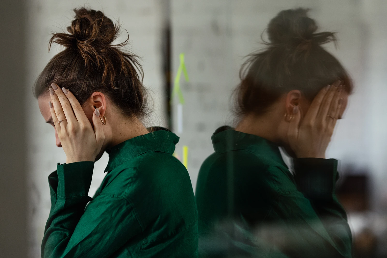 A young woman with her hair in a bun covers her face with her hands in a moment of deep sadness or stress in an office.