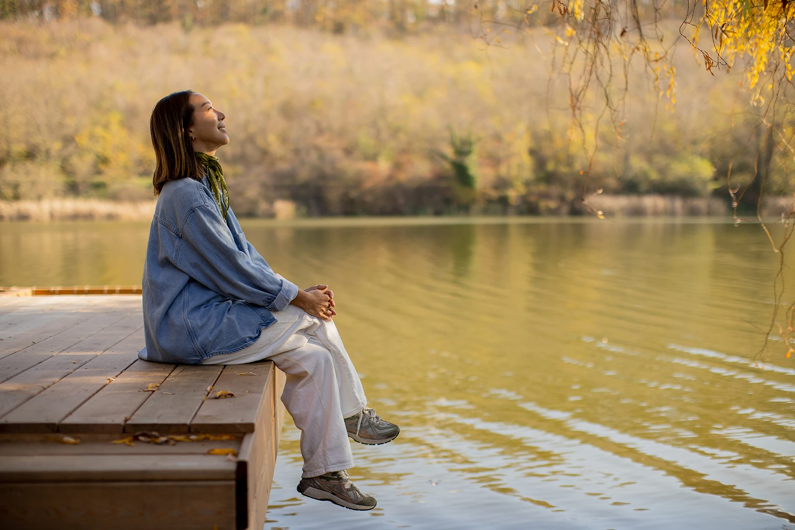 A relaxed woman sits peacefully on a wooden dock by a calm lake, smiling with her eyes closed while enjoying a scenic autumn nature view.